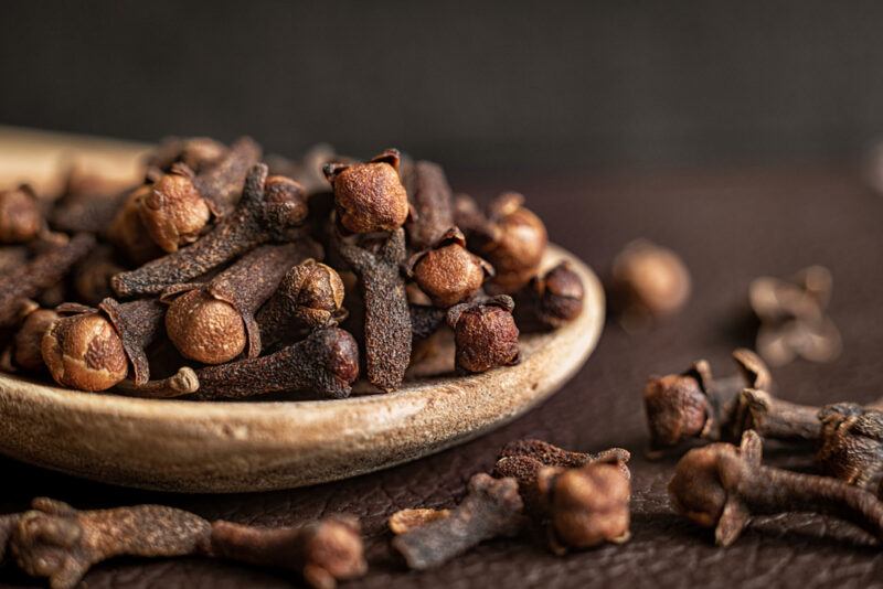 on a dark brown surface is a closeup image of a wooden ladle full of cloves with loose cloves around it