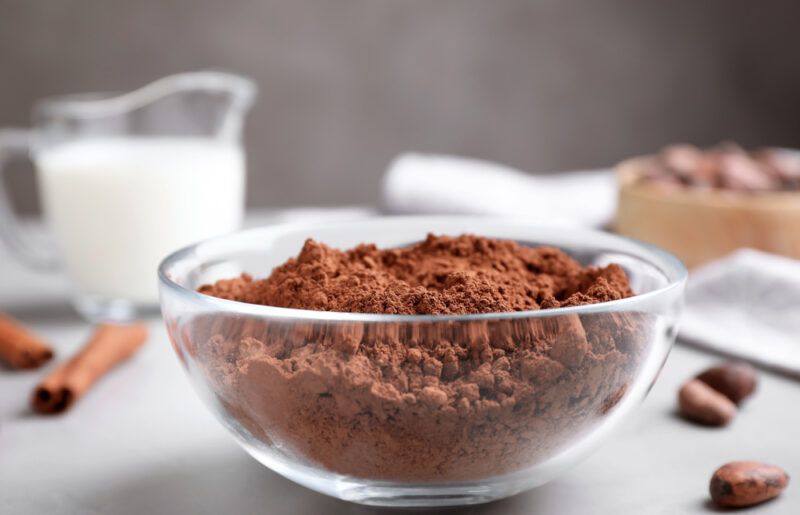 a closeup image of a clear glass bowl with cocoa butter, at the back is a small pitcher of milk, a couple of cinnamon sticks, and a few loose nuts