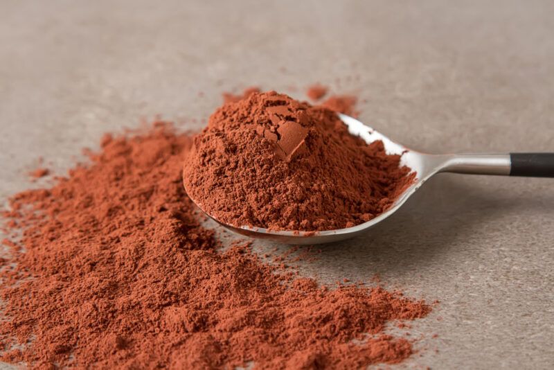 closeup image of a spoonful of cocoa powder resting on a concrete surface with a small mound of cocoa powder beside it