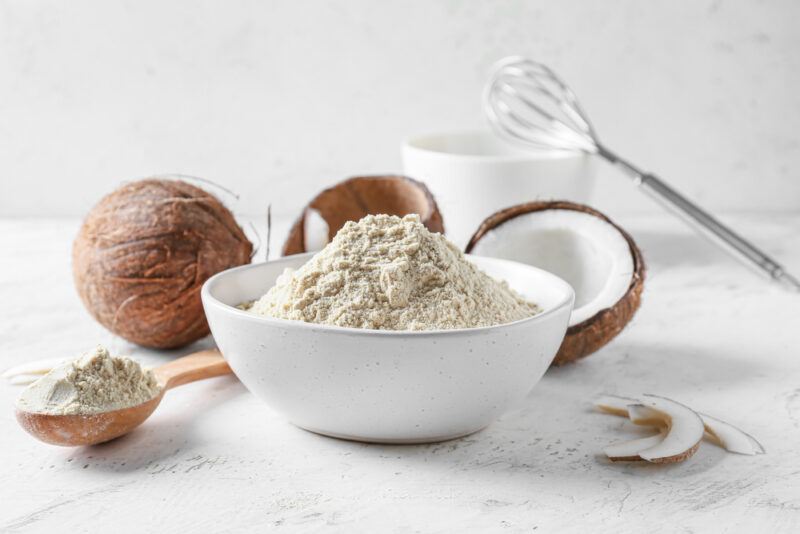 on a marble surface is a white ceramic bowl full of coconut flour with a wooden spoon beside it full of coconut flour, at the back is a whole coconut and halve coconut, a white bowl with a metal whisk on it