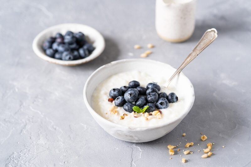 on a grey concrete surface is a white bowl of coconut porridge with fresh blueberries, nuts, and a sprig of herb on top, around it are loose crushed nuts, at the back is a small white dish with fresh blueberries and a small white jar of coconut milk