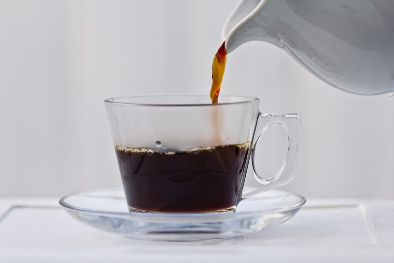 a clear cup and saucer with black coffee being poured from a white ceramic pitcher