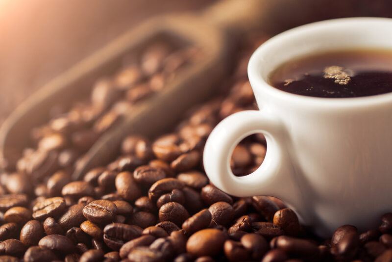 a closeup image of a mound of coffee beans with a wooden scoop shoved in it, selective focus on a partial image of a white cup of coffee