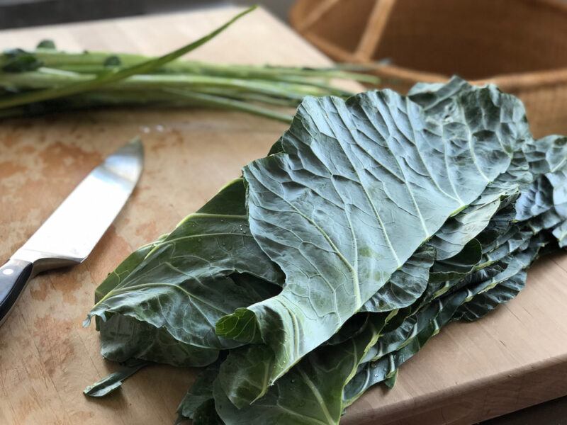 on a wooden chopping board are cut collard greens with a knife beside it, the cut out thick center rib of the leaves can be seen at the back 