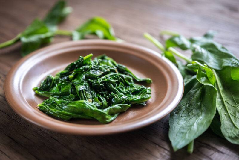 on a wooden surface is a brown clay dish with cooked spinach, around it are fresh spinach leaves