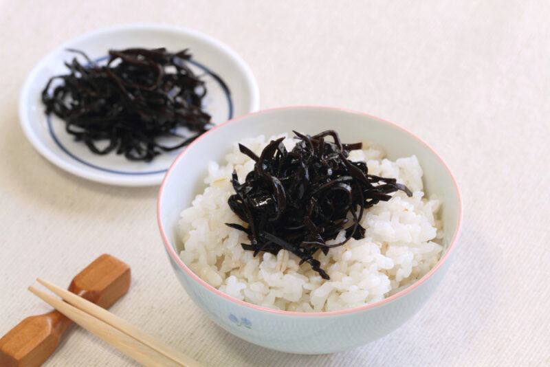 cooked arame on top of steamed rice in a white bowl with chopsticks beside it, at the back is a small dish with arame