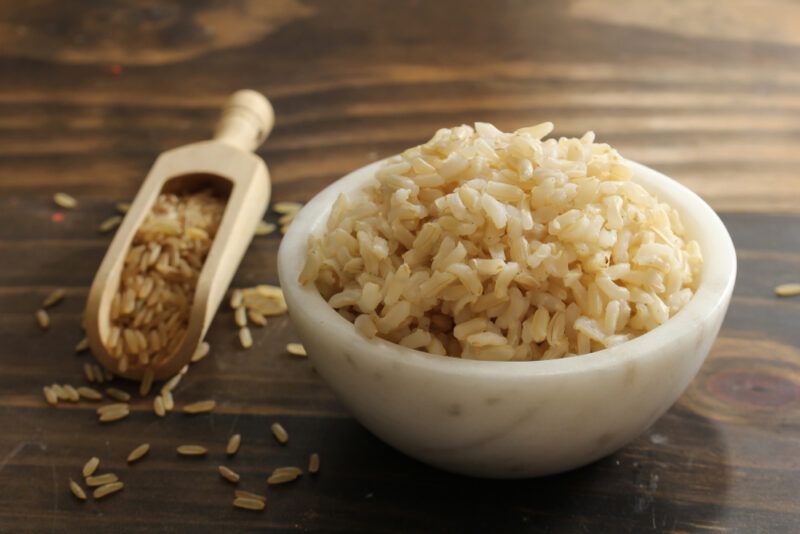 on a dark wooden surface is a white marble bowl full of cooked brown rice, beside is a wooden scoop full of raw brown rice with loose grains around it