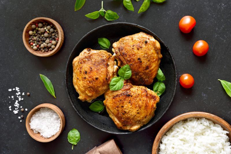 three cooked chicken breasts topped with basil on a black bowl, a bowl of peppers and salt, and a bowl of rice on a dark background