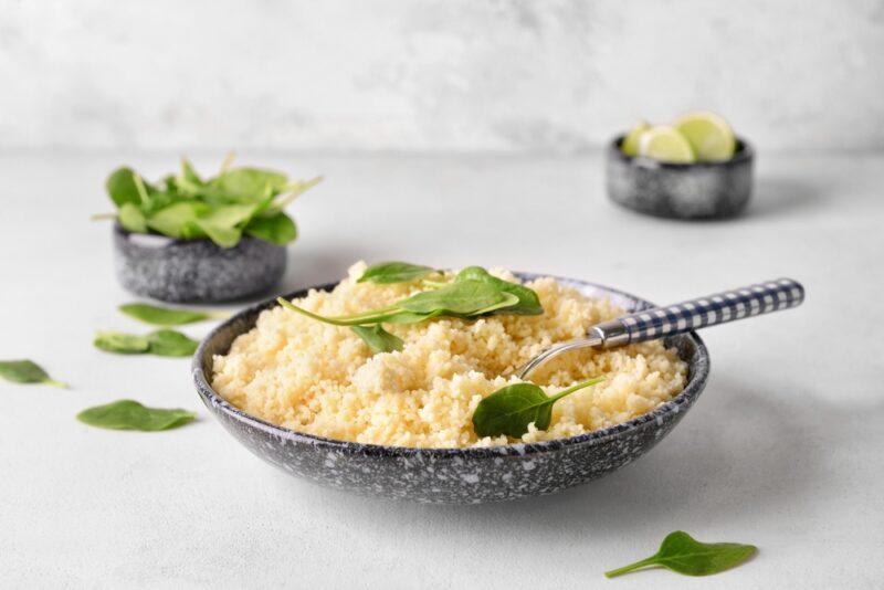 on a white surface is a stone shallow bowl full of boiled couscous with herbs on top, around it are loose herbs, at the back is a couple of small stone bowls, one with fresh herbs, the other with sliced limes