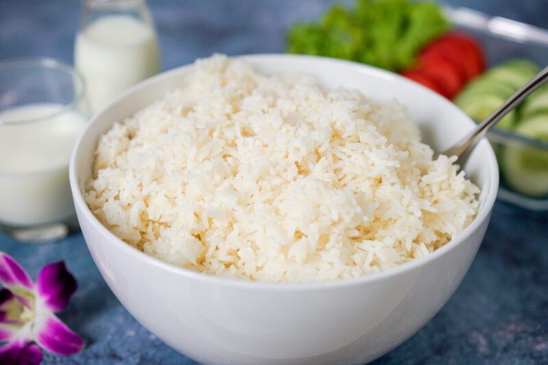 closeup image of a white bowl full of cooked white rice with fresh vegetables at the back and a couple of glasses of milk