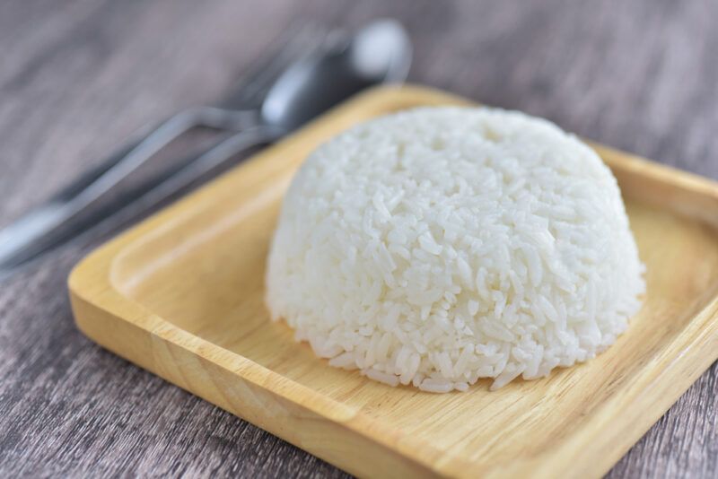 on a rustic looking wooden surface is a square wooden dish with a molded cup of cooked white rice, beside it is a silver spoon and fork