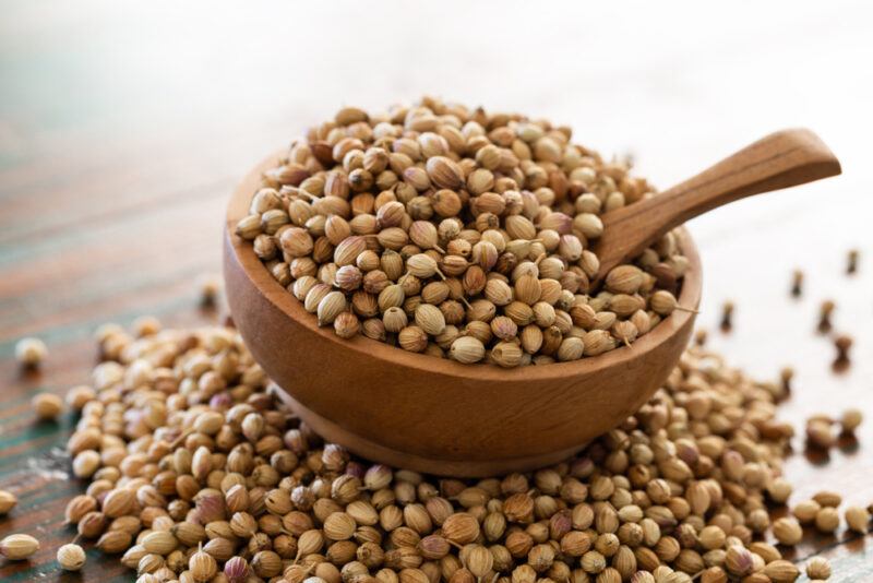 on a wooden surface is a mound of coriander seeds with a small wooden bowl full of coriander seeds and a wooden spoon