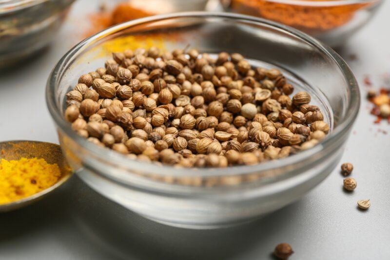 a closeup image of a glass bowl full of coriander seeds, around it are loose coriander seeds and small containers with different ground spices