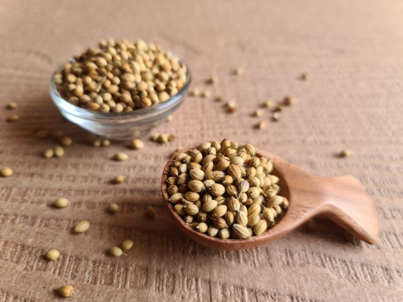 on a wooden surface is a short wooden spoon with coriander seeds, and small clear bowl with coriander seeds, around it are loose coriander seeds