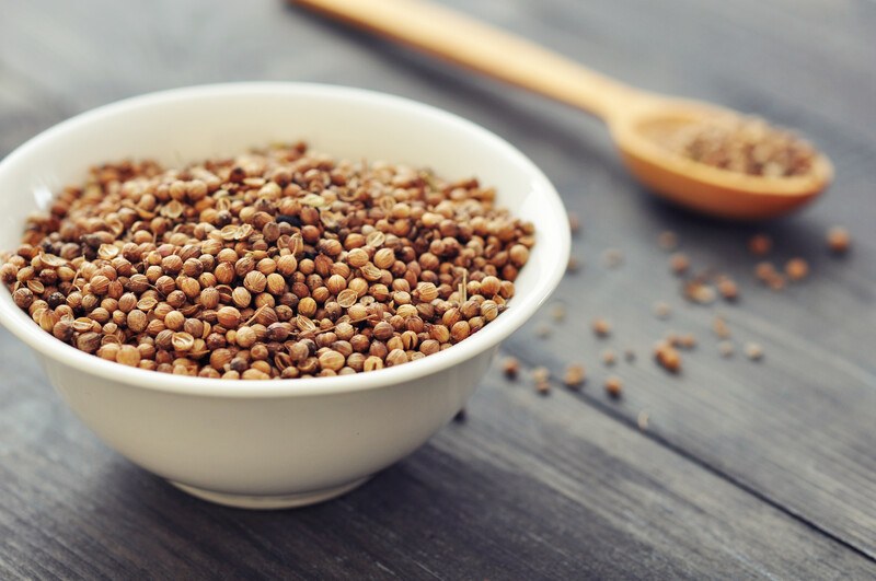 closeup image of a white bowl full of coriander seeds resting on a wooden surface with loose coriander seeds and wooden spoon full of coriander seeds at the back