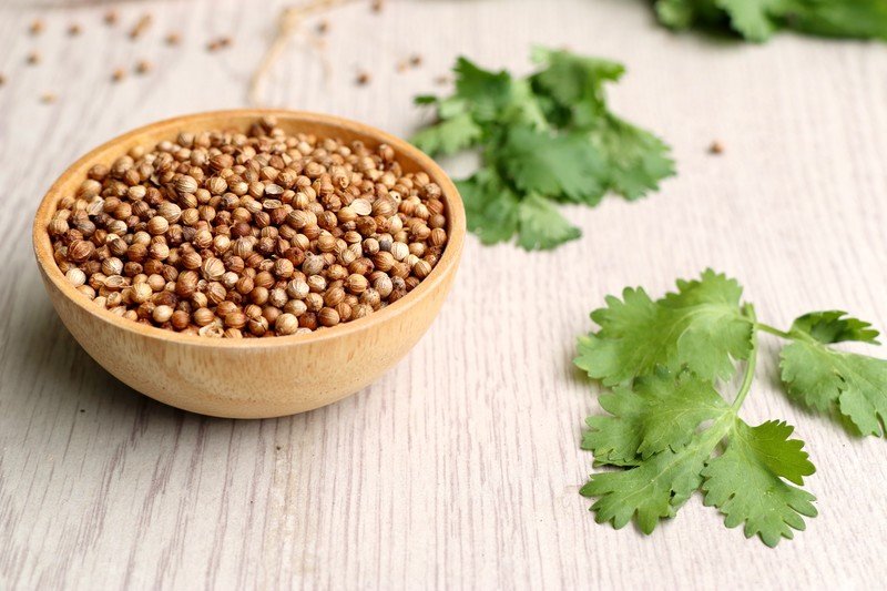 a wooden bowl full of coriander seeds resting on a wooden surface with loose coriander seeds and fresh coriander leaves around it