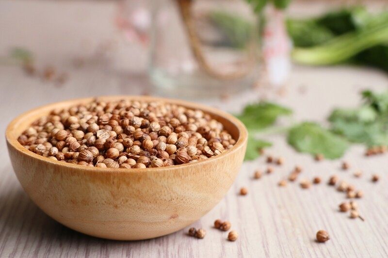 on a wooden surface is a wooden bowl full of coriander seeds with loose coriander seeds and fresh leaves around it 