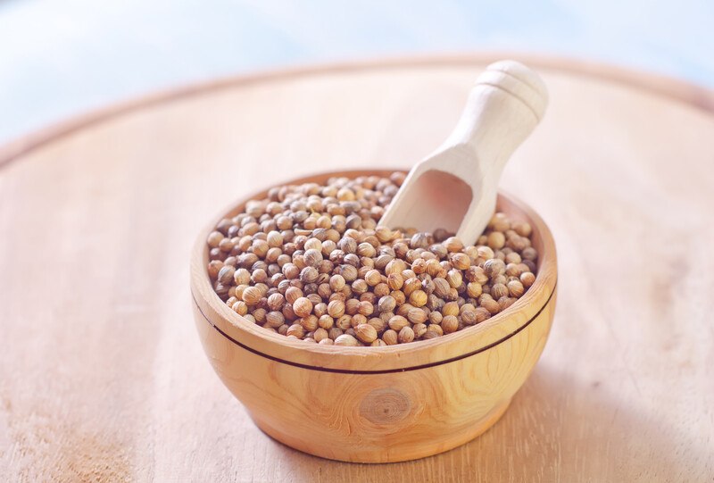 on a wooden round board is a wooden bowl full of coriander seeds with a wooden scoop 