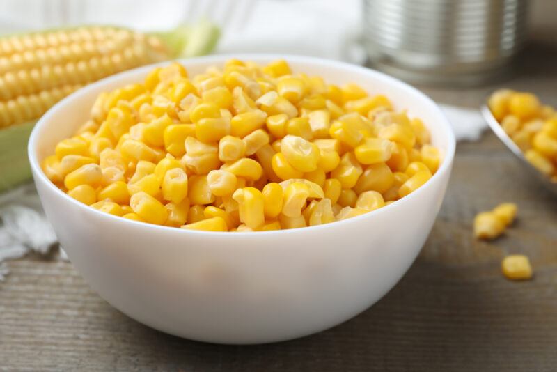 on a wooden surface is a white bowl full of corn kernels with loose corn kernels and ear of corn at the back