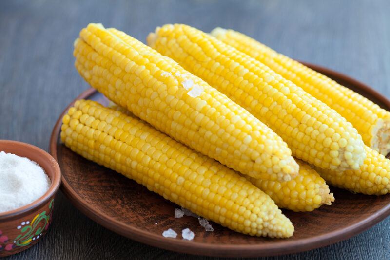 closeup image of a brown wooden plate full of boiled corn on the cobs sprinkled with rock salt, beside it is a small wooden bowl of rock salt