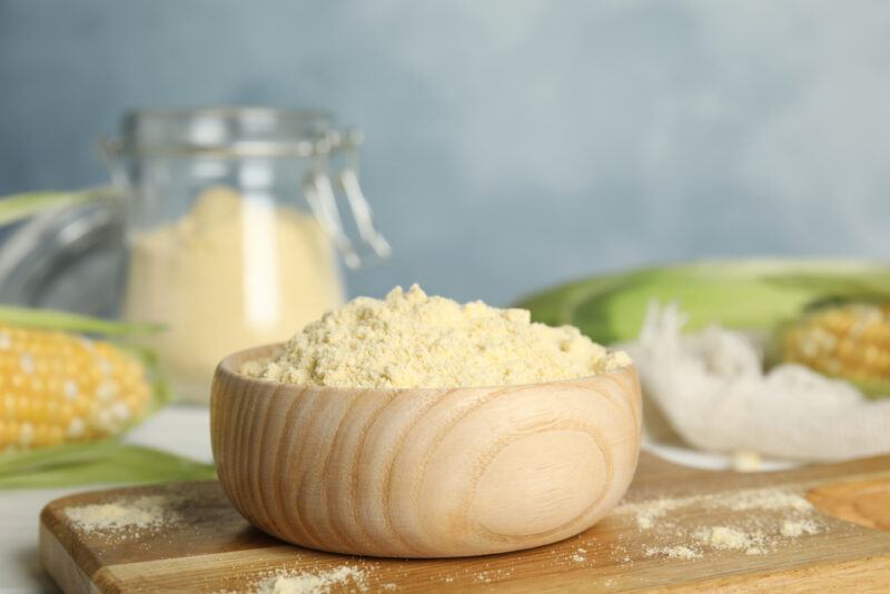 a light colored wooden bowl full of corn flour, resting on top of a wooden chopping board, behind it are a couple of ear of corn and a jar of corn flour