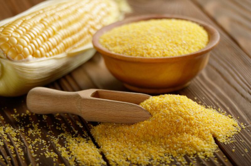 on a rustic looking wooden surface is a bowl of corn grits with a small mound in front of it with a wooden scoop, at the back is a corn on the cob