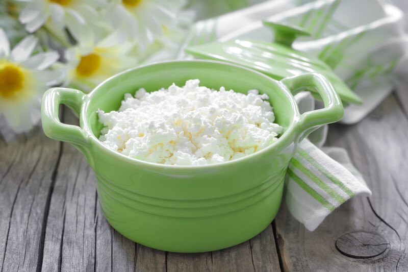 on an aged wooden surface is a green bowl with handle full of cottage cheese, with white flowers and white and green table napkin at the back