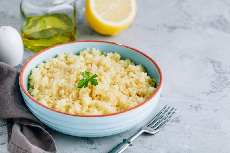 on a marble surface is a blue bowl with orange rim full of cooked couscous with a tiny  cilantro leaf on top, at the back is a halve lemon, jar of olive oil and pepper shaker, beside it is silver fork and a dark-colored table napkiin