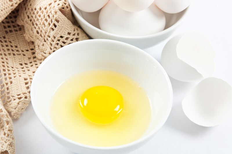 on a white surface is a white bowl with cracked egg with the yolk and egg white, at the back is a brown knitted textile and a bowl of fresh white eggs