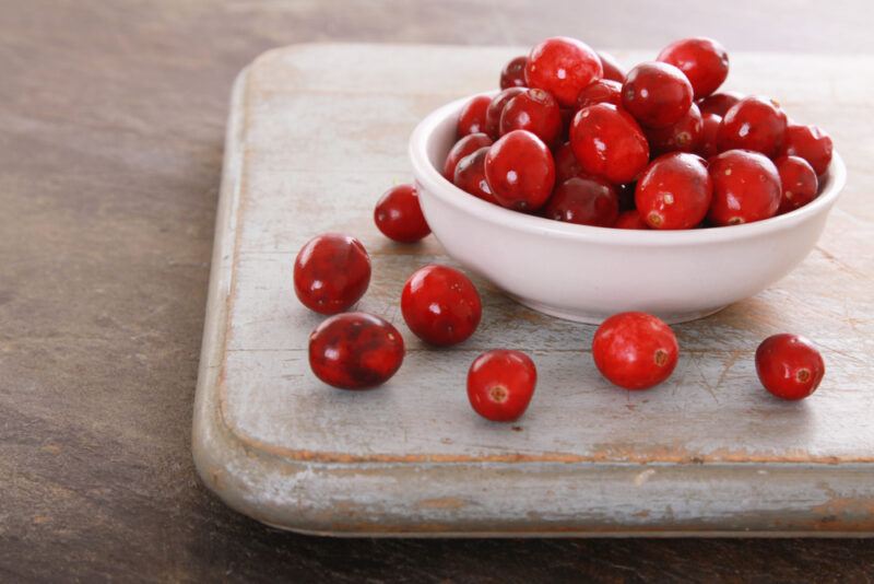 on dark concrete surface is a rustic looking wooden chopping board is a small white dish full of cranberries with loose cranberries around it