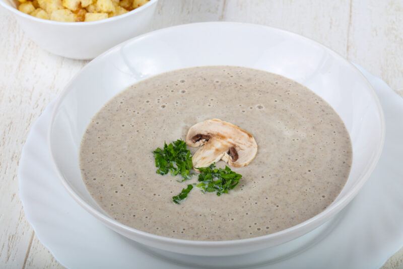 a white bowl with cream of mushroom soup with a sliced mushroom and sprig of herb on top