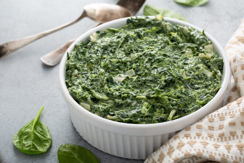 on a concrete surface is a ramekin full of creamed spinach, around it is a a a couple of fresh spinach, silver spoons, and cream-colored kitchen towel with pattern