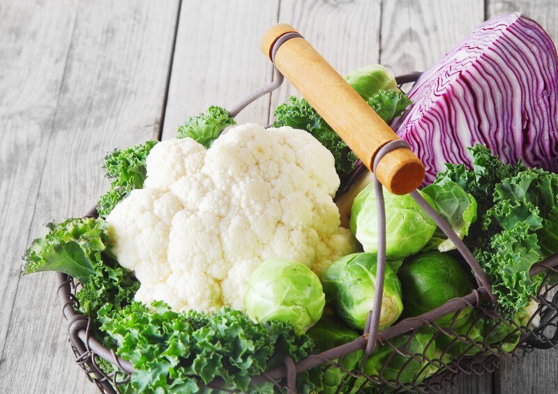 on an aged wooden surface is a wire basket with wooden handle full of cruciferous vegetables