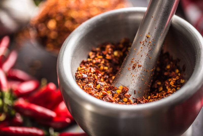 metal mortar and pestle with crushed red pepper, beside it are fresh chili peppers, and a bowl of crush red peppers