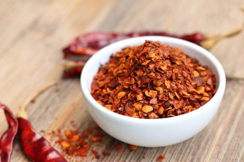 on a wooden surface if a white bowl with crushed red pepper, around it are loose crushed red pepper and dried red chilis