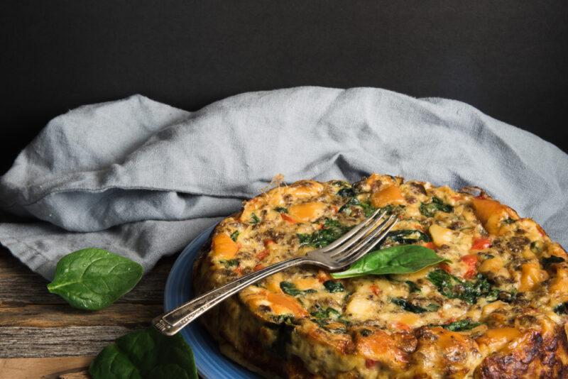 on a wooden surface is a blue plate with a crustless spinach, mushroom, and feta quiche with a single fresh spinach on top and a couple more beside it, at the back is a grey-colored table napkin