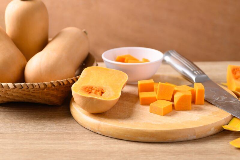 on a wooden surface is a wooden chopping board with cubed butternut squash, beside it is a halved butternut squash, knife, and white bowl with cubed butternut squash, at the back is a weaved tray with whole butternut squash