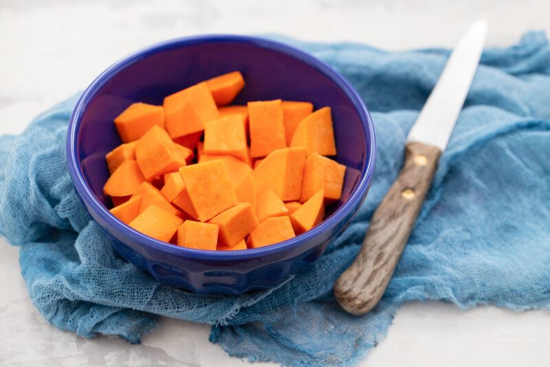 a dark blue ceramic bowl full of cubed sweet potatoes resting on top of a blue cloth with a knife beside it