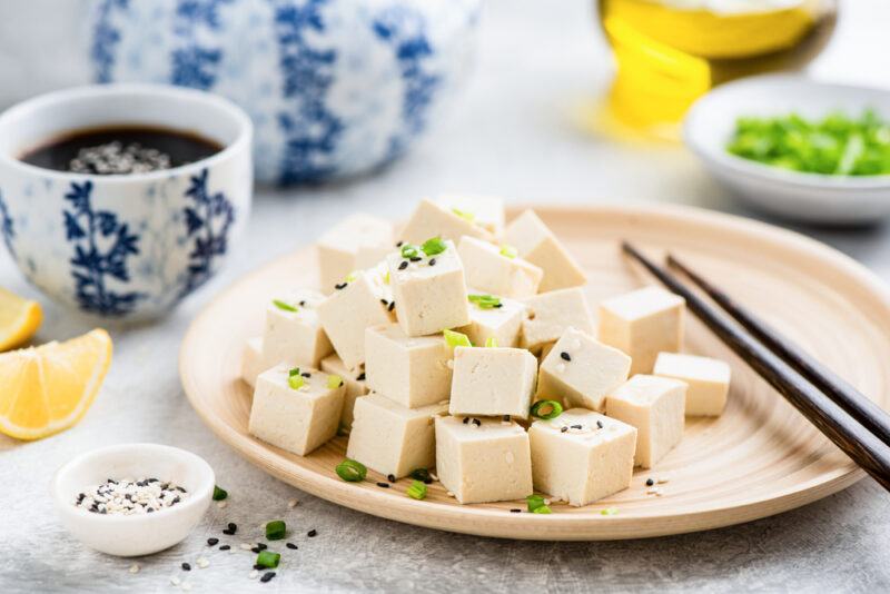 a beige wooden plate of cubed tofu with minced chives and white and black sesame sprinkled on top with chopsticks beside it, around the plate are small white dishes of white and black sesame seeds, minced chives, soy sauce, olive oil, and lemon wedges