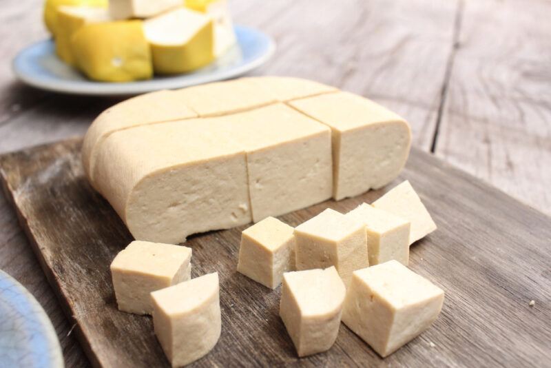 on a rustic looking wooden surface is an aged wooden surface with cubed tofu on it, at the back is a bluish saucer with cube tofu as well