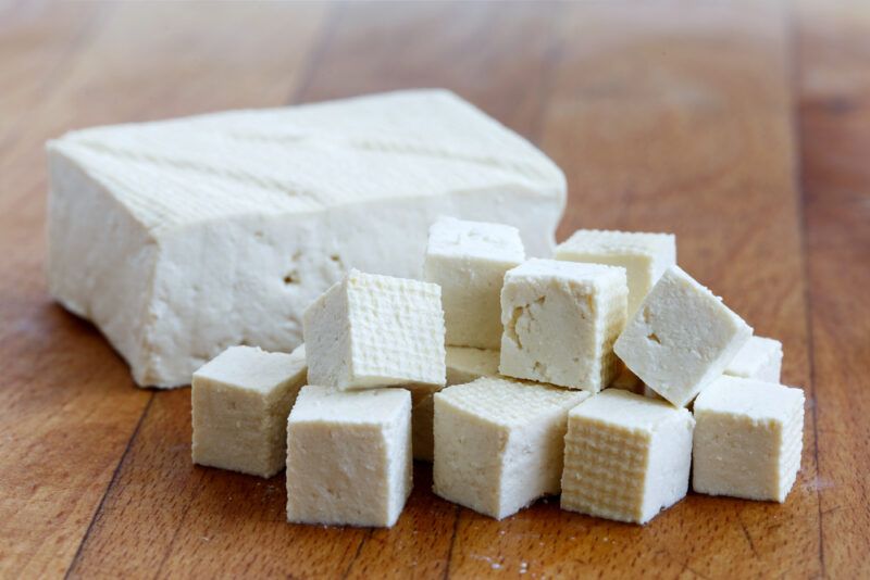 on a wooden surface is a pile of cubed tofu, behind is a block of fresh tofu