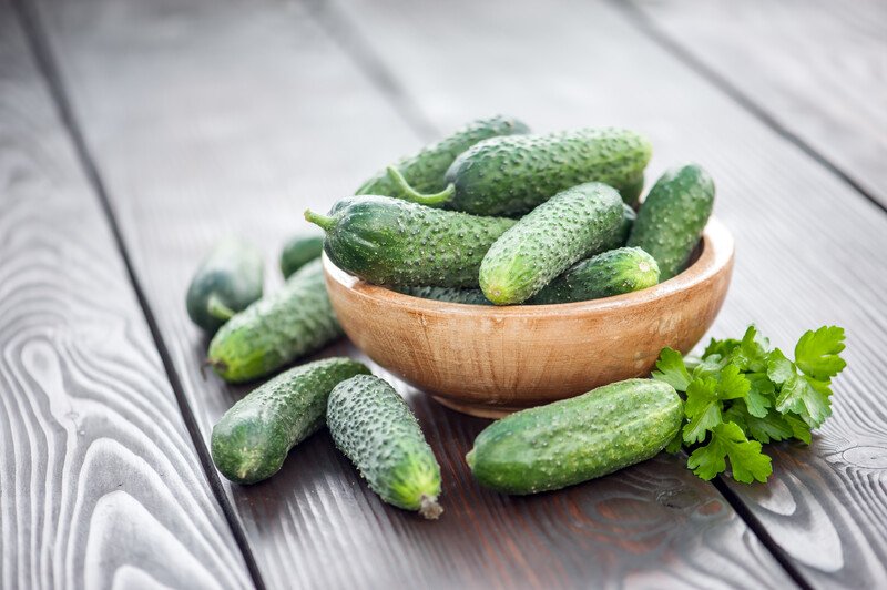 on a dark wooden surface is a wooden bowl of cucumber with cucumber around it a sprigs of parsley