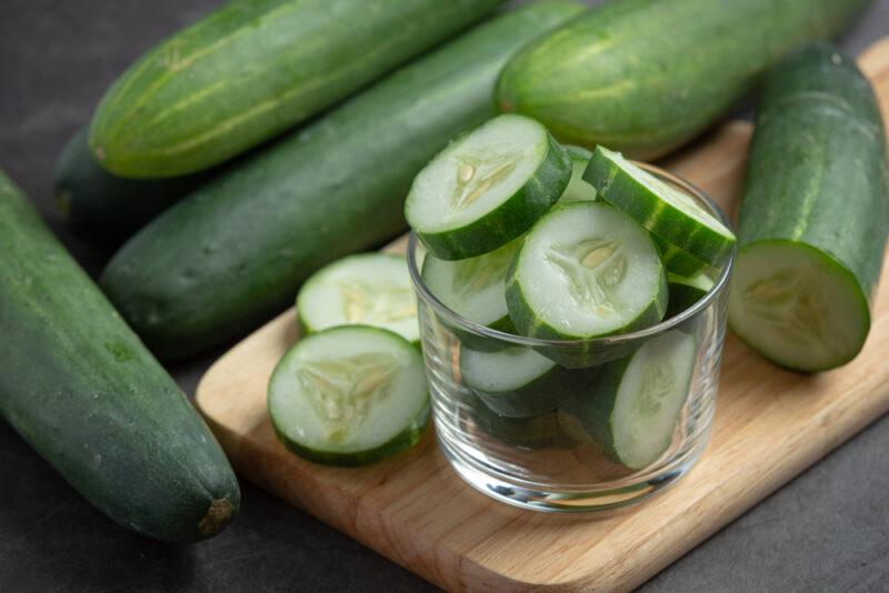 a closeup image of sliced cucumber resting on top of a wooden chopping board with whole zucchinis behind it