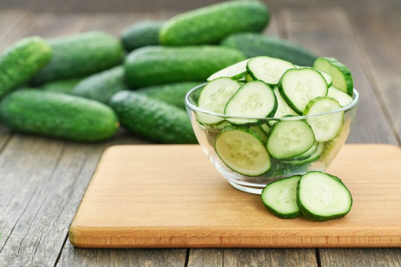 on a wooden surface is a wooden chopping board with a clear glass bowl of sliced cucumber, at the back is a pile of fresh cucumbers