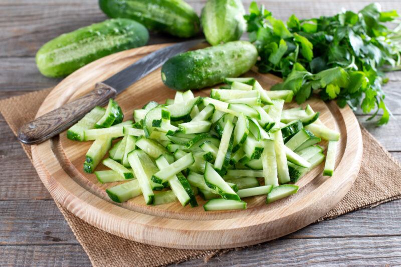 on an aged wooden surface is a round wooden board with chopped cucumber sticks, with a knife beside it and a whole cucumber, resting on top of a square burlap, with whole cucumbers and cilantro herbs at the back