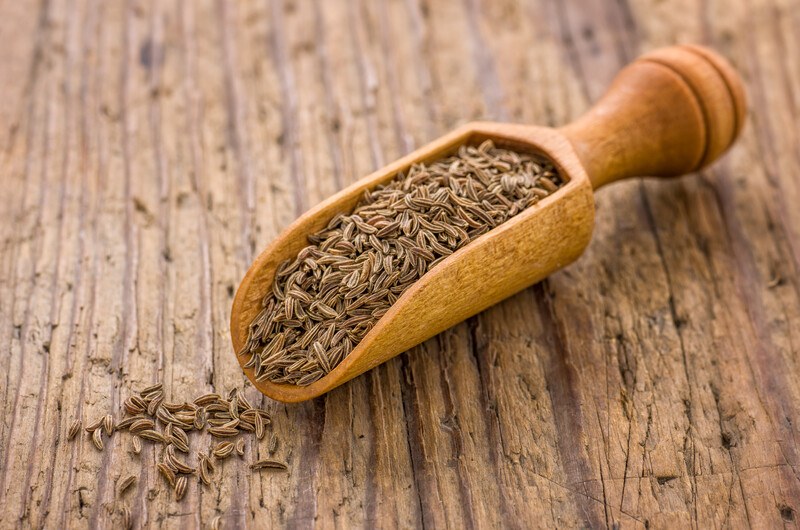 a wooden scoop full of cumin seeds resting on an aged-wooden surface