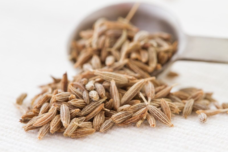 on a white table napkin is a closeup image of cumin seeds pouring out of a metal measuring spoon