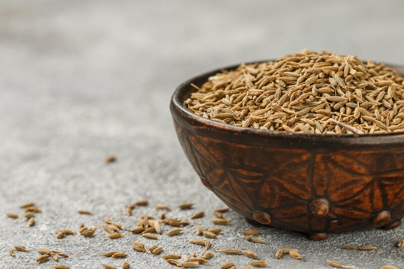 closeup image of a dark brown textured bowl full of cumin seeds, with loose cumin seeds around it.