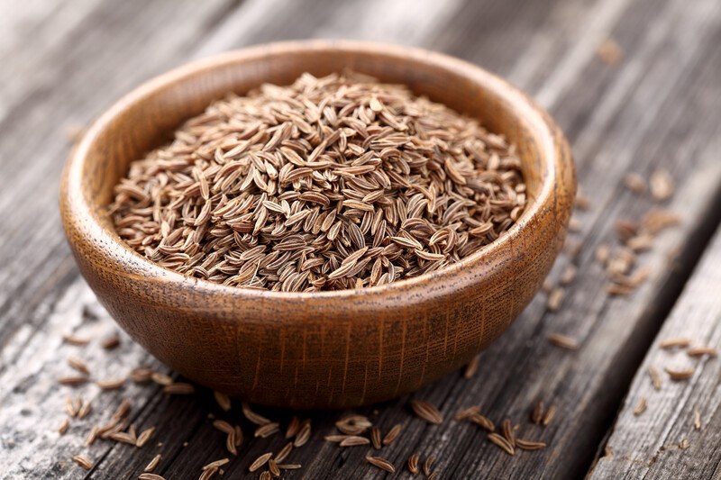 on a dark and aged wooden surface is a brown wooden bowl full of cumin seeds, with loose cumin seeds around it