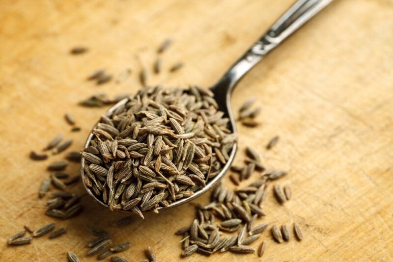 on a wooden surface is a silver spoon full of cumin seeds, with loose cumin seeds around it